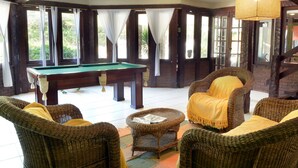 Una mesa de billar en una habitación con muebles de mimbre y ventanas amplias.