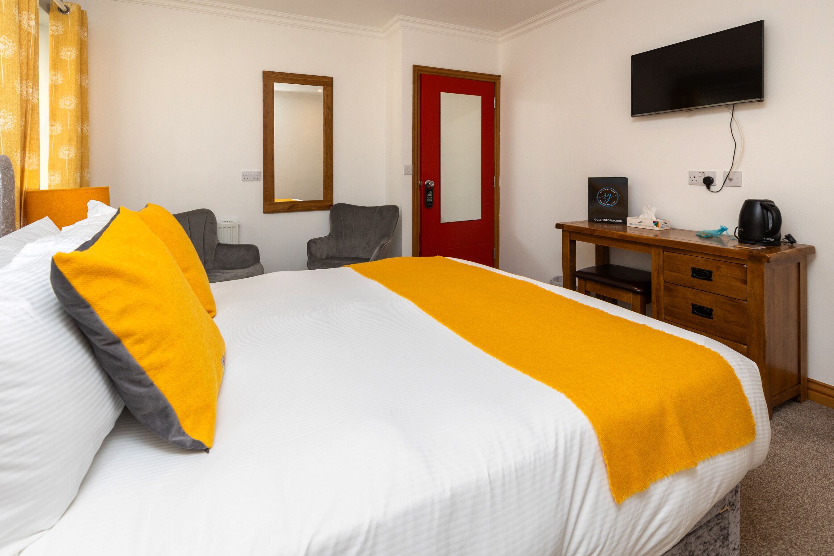 A hotel room with a bed, a desk, a chair, a television, and a red door.