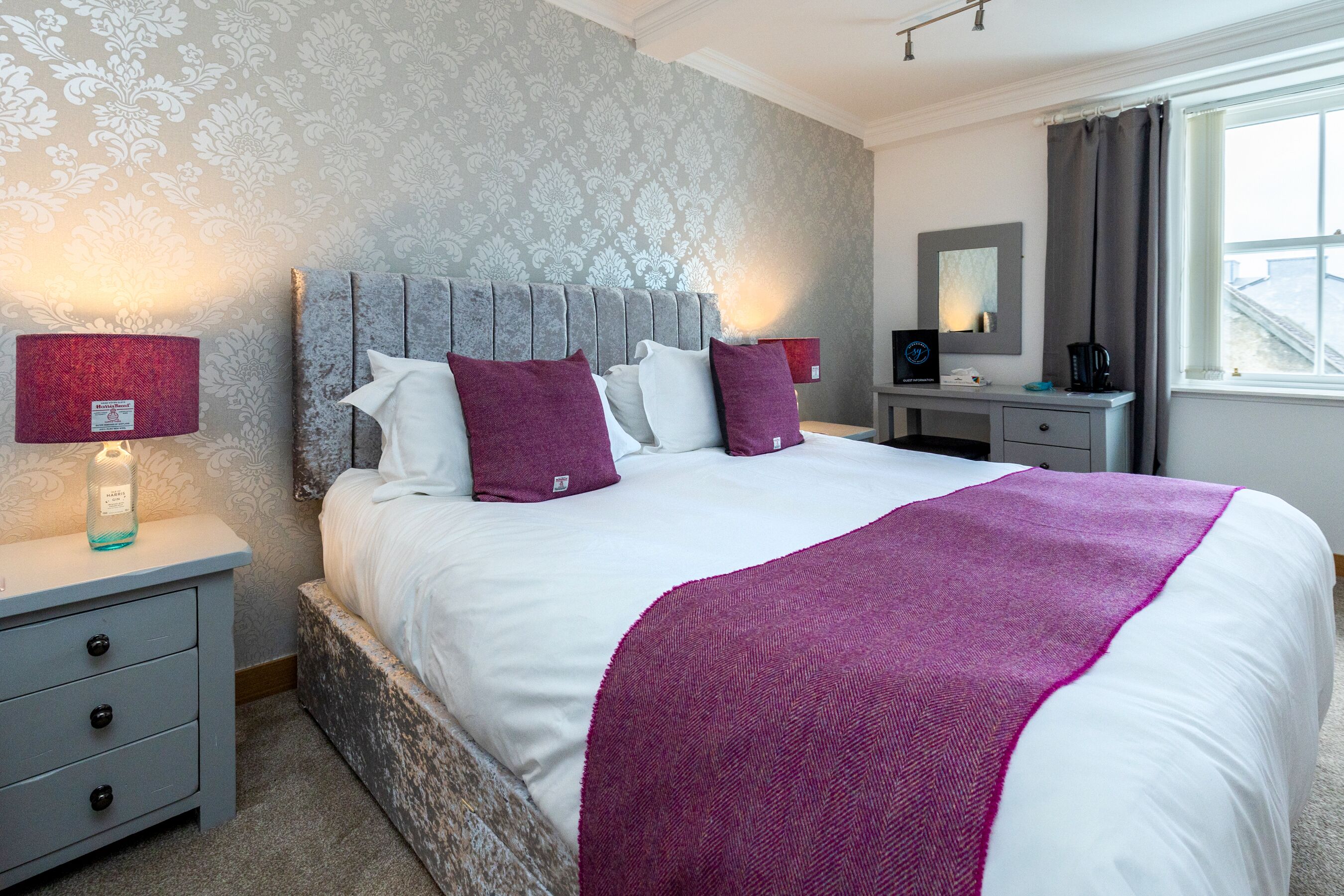 A neatly made bed with white and purple bedding, a grey headboard, and a bedside table with a lamp and a bottle.