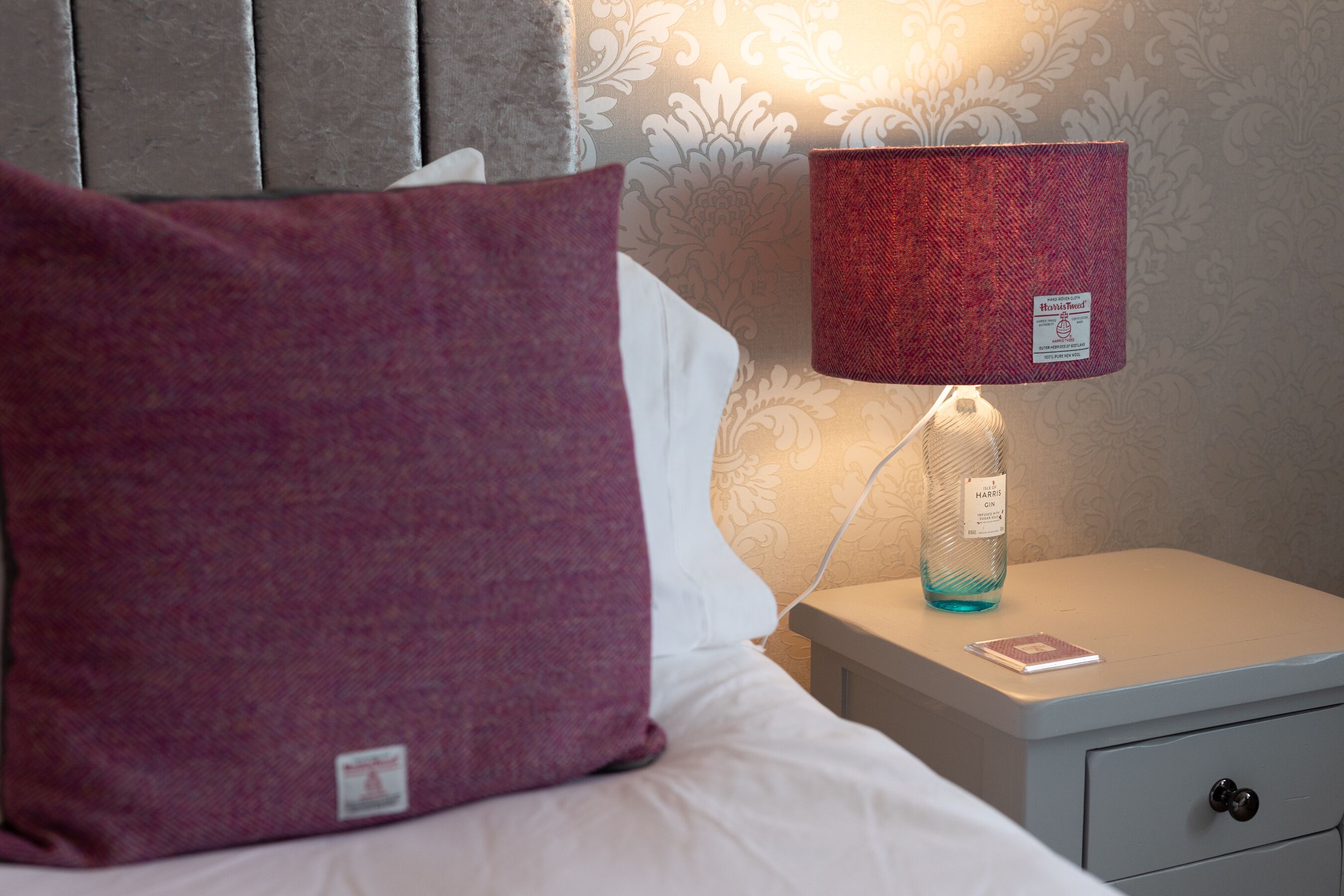 Ein ordentlich bezogenes Bett mit einem großen violetten Kopfkissen, einem weißen Kopfkissen und einer roten Lampe auf einem Nachttisch.
