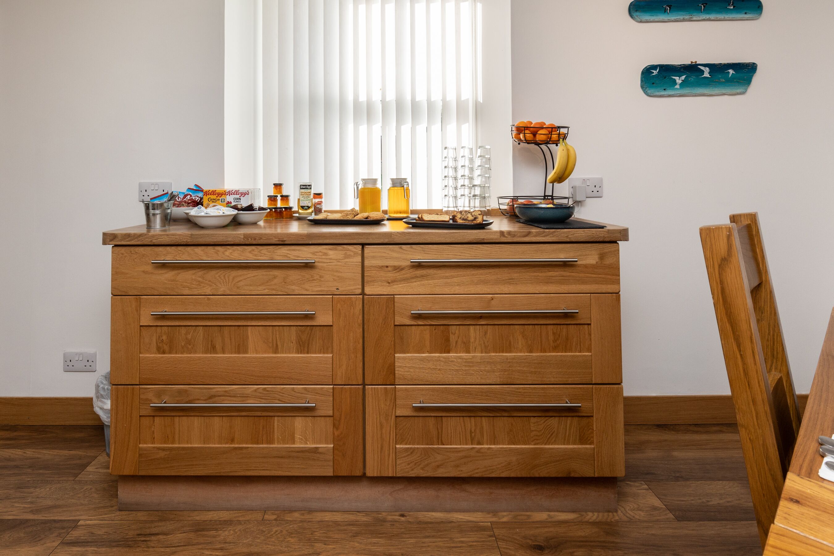 Un comptoir de cuisine en bois, sur lequel sont disposés divers aliments pour le petit-déjeuner, une fenêtre avec des stores et des œuvres d’art décoratives accrochées au mur.