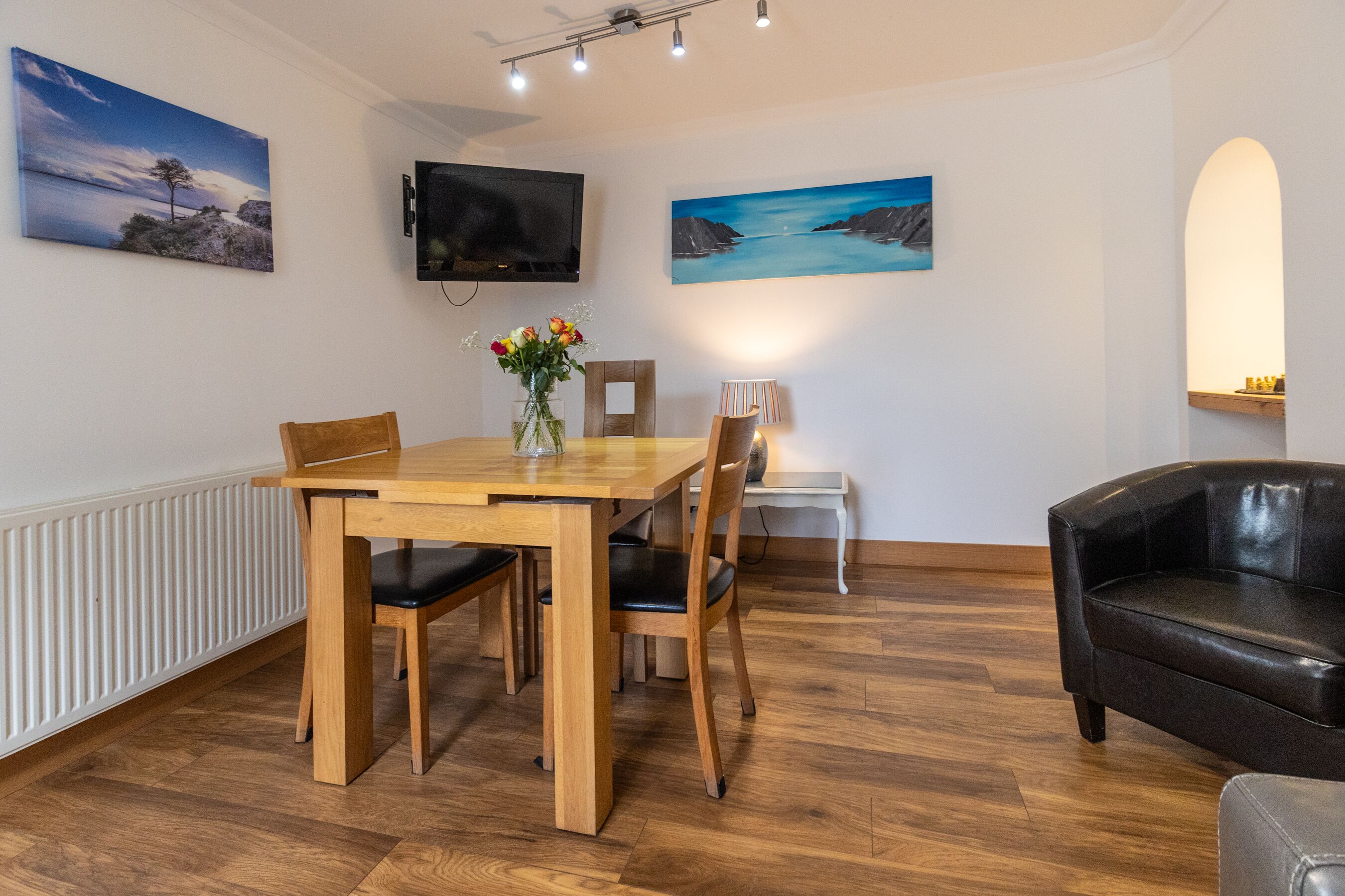 A dining area with a wooden table and chairs, a vase with flowers, a television on the wall, and artwork depicting coastal scenes.