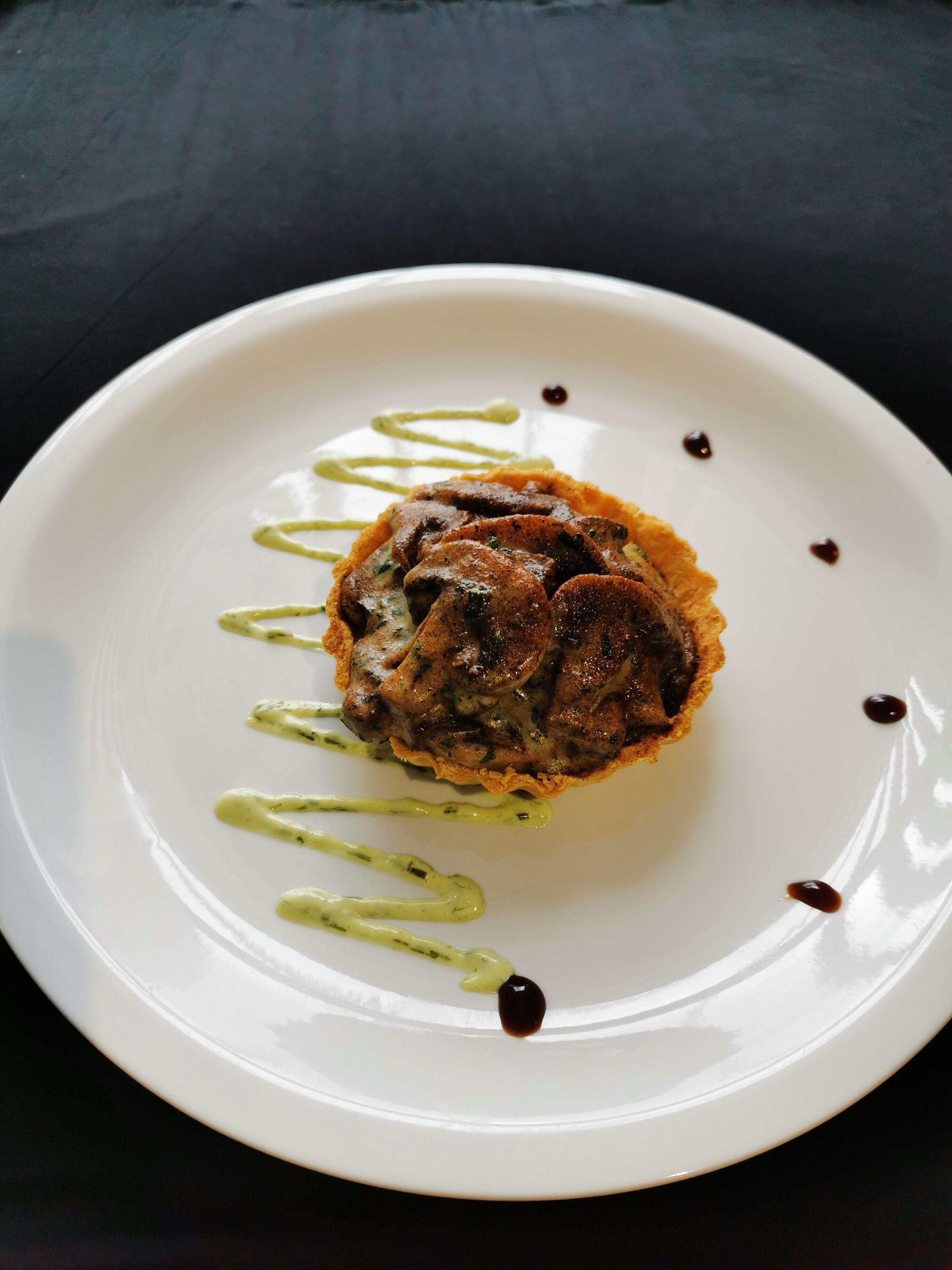 A white plate with a food item, possibly a meat-based dish, garnished with green sauce and small dark dots.