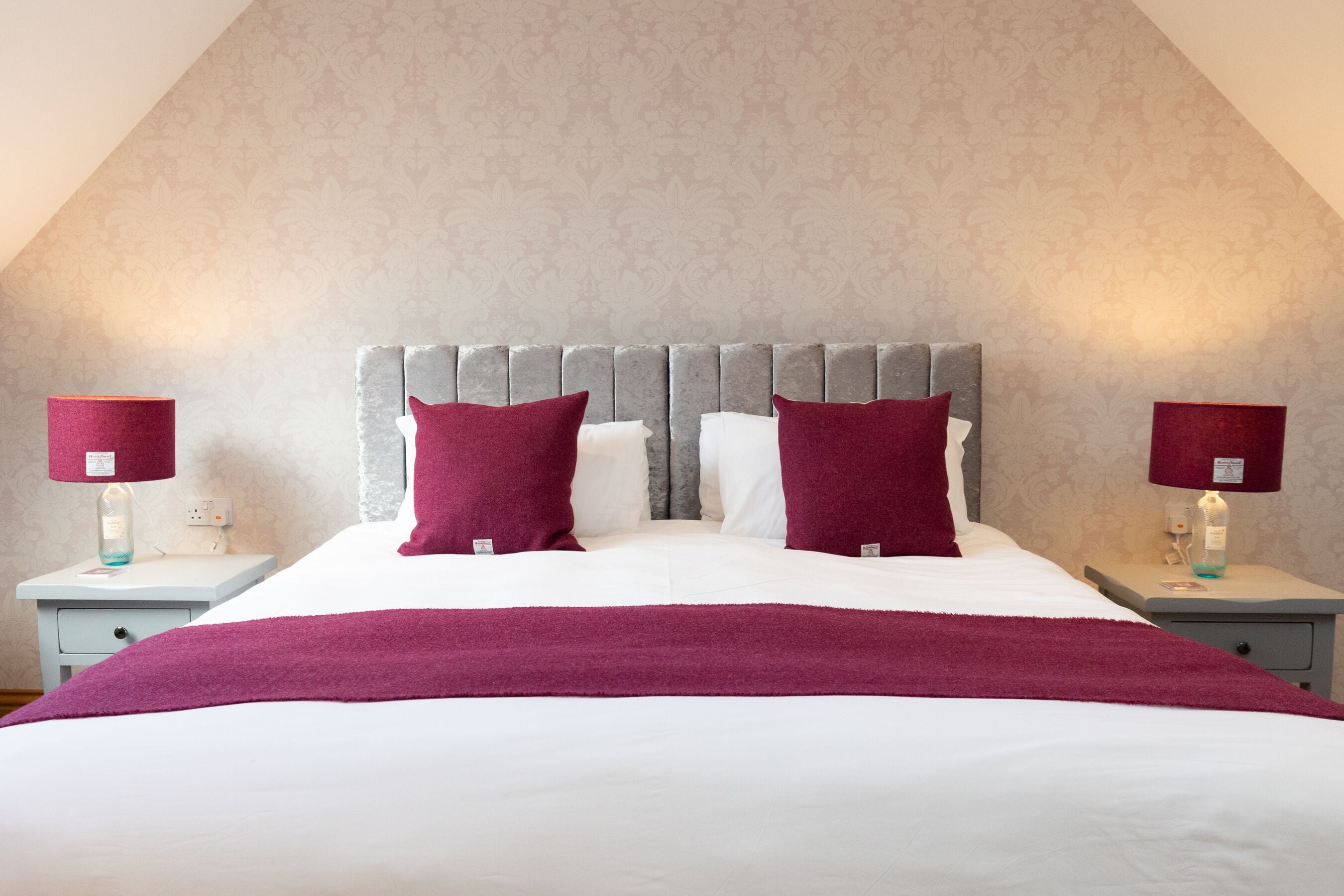 A neatly made bed with a grey upholstered headboard, two red pillows, and a white comforter. There are two bedside tables with lamps and a small drawer.