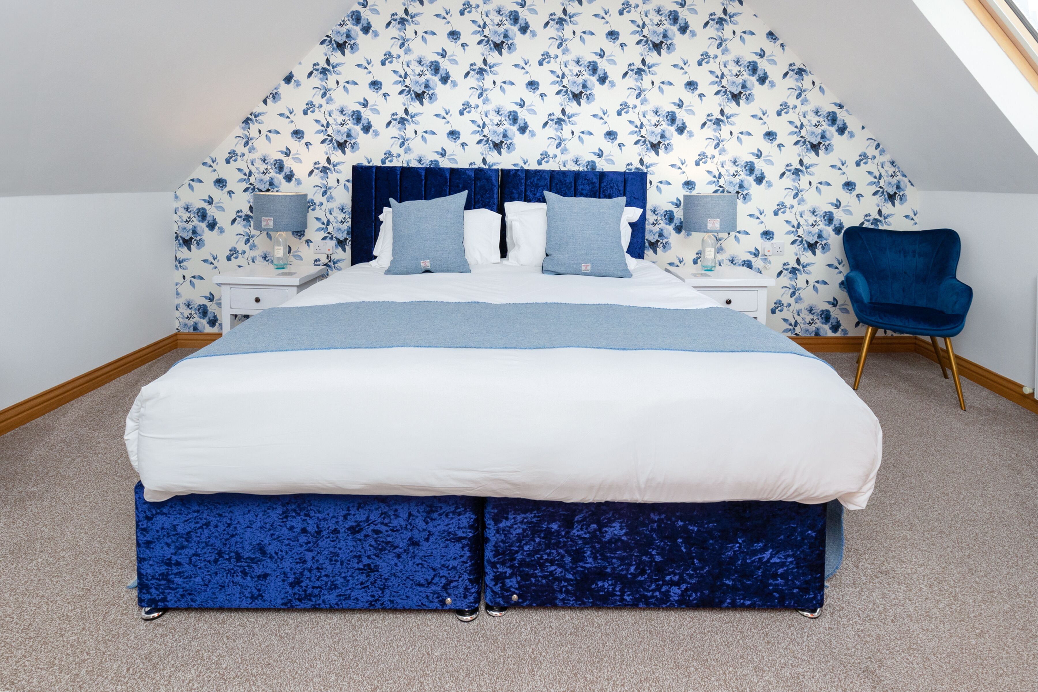 Une chambre avec un grand lit, un papier peint à motifs floraux, une chaise bleue et une lucarne.