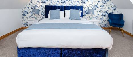 Une chambre avec un grand lit, un papier peint à motifs floraux, une chaise bleue et une lucarne.