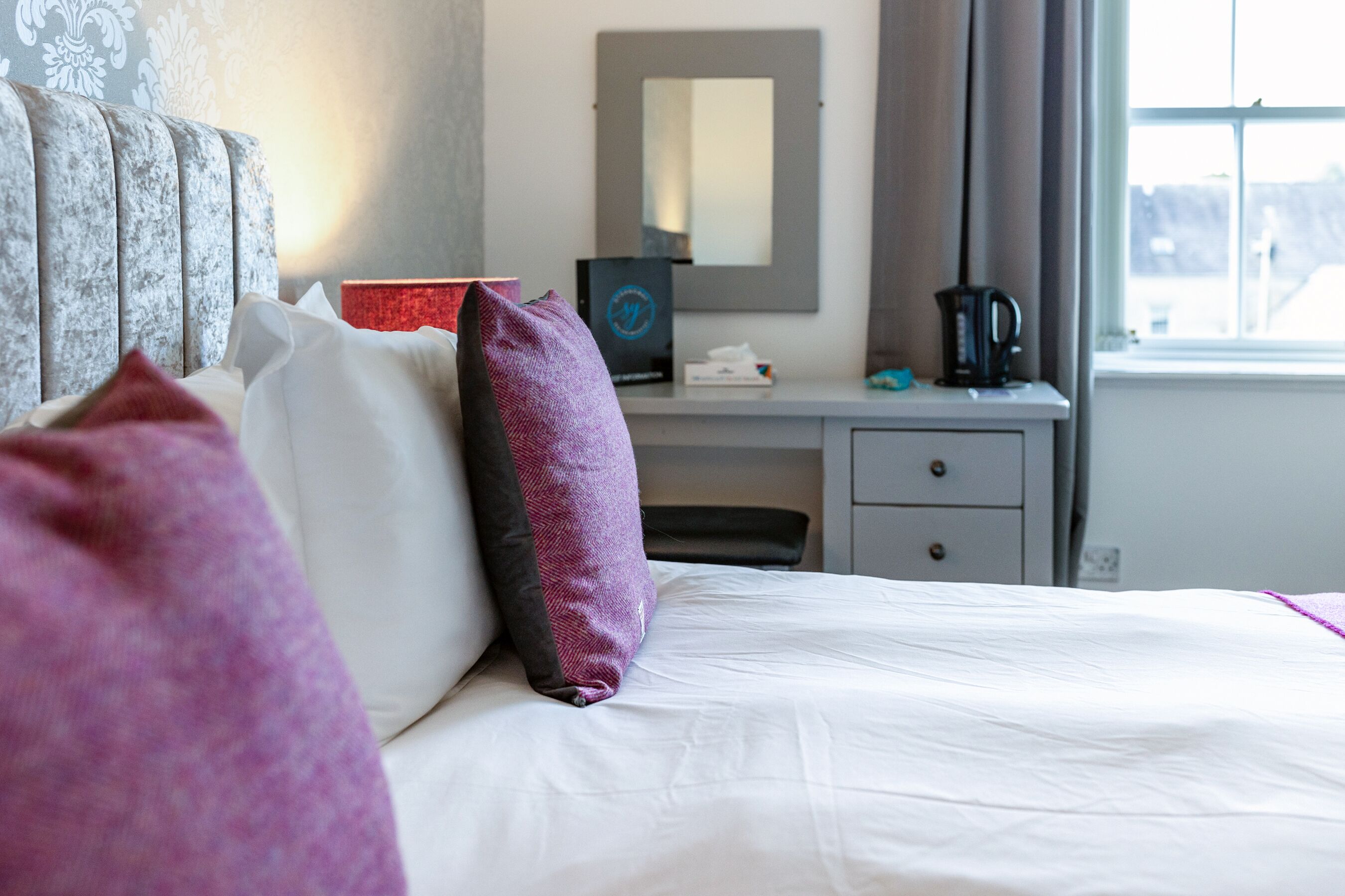 A hotel room with a bed, pillows, a desk with a mirror, a coffee maker, and a window with a view.