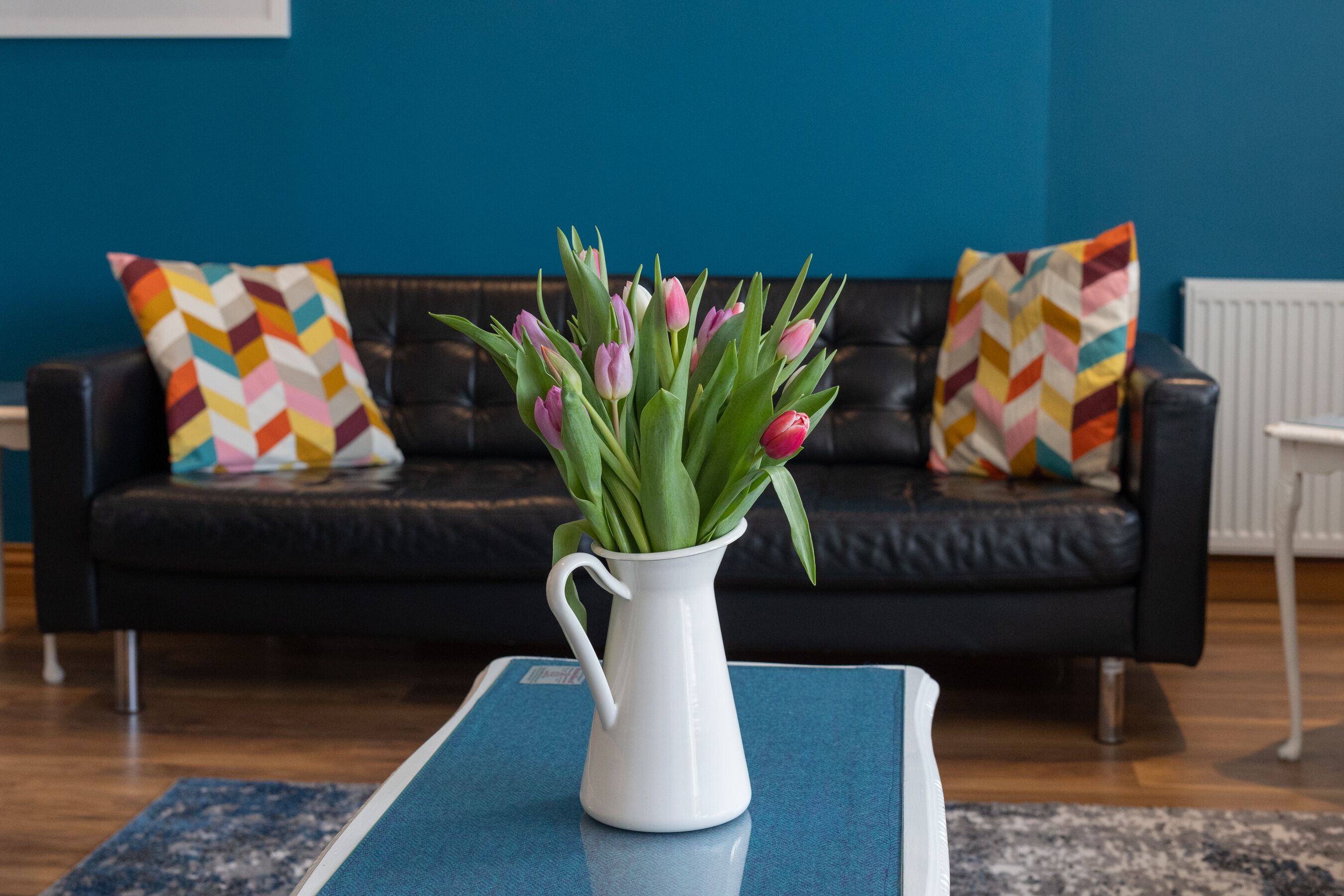 A white vase with pink tulips on a blue table in a living room with a black leather couch and colorful patterned pillows.