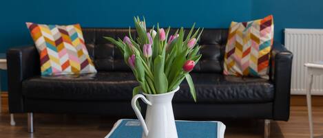 A white vase with pink tulips on a blue table in a living room with a black leather couch and colorful patterned pillows.