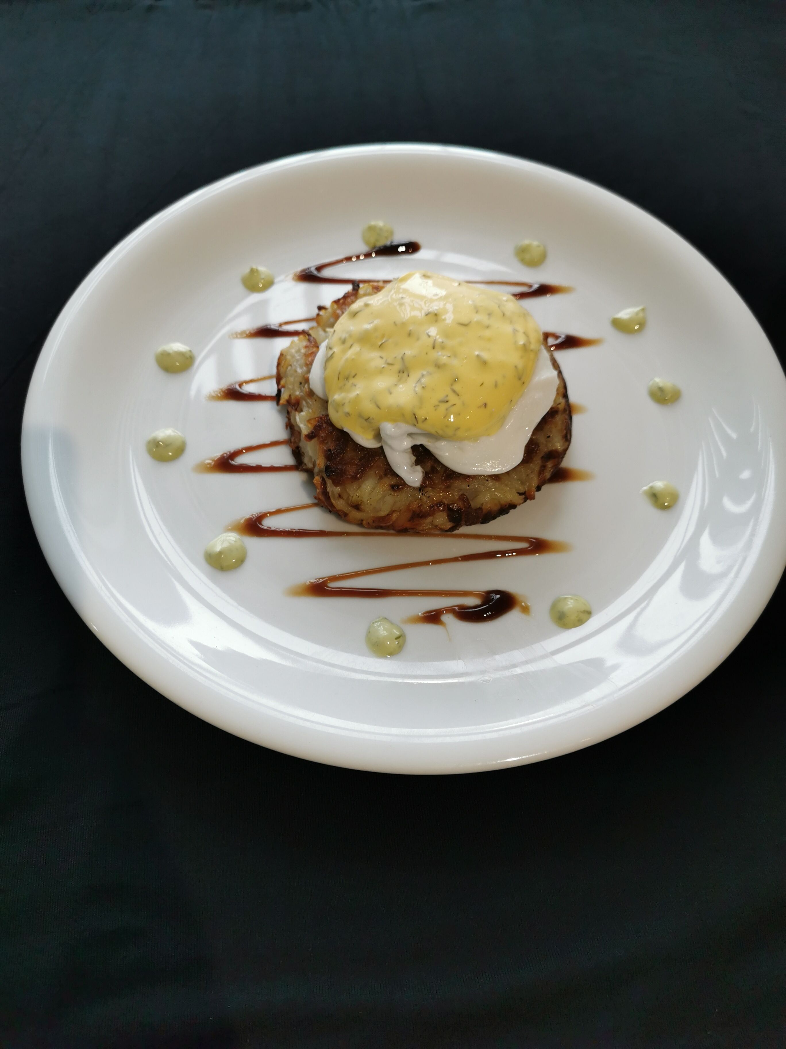 Un plato blanco con un alimento cubierto de salsa cremosa y rociado con salsa marrón, rodeado de pequeños puntos verdes.