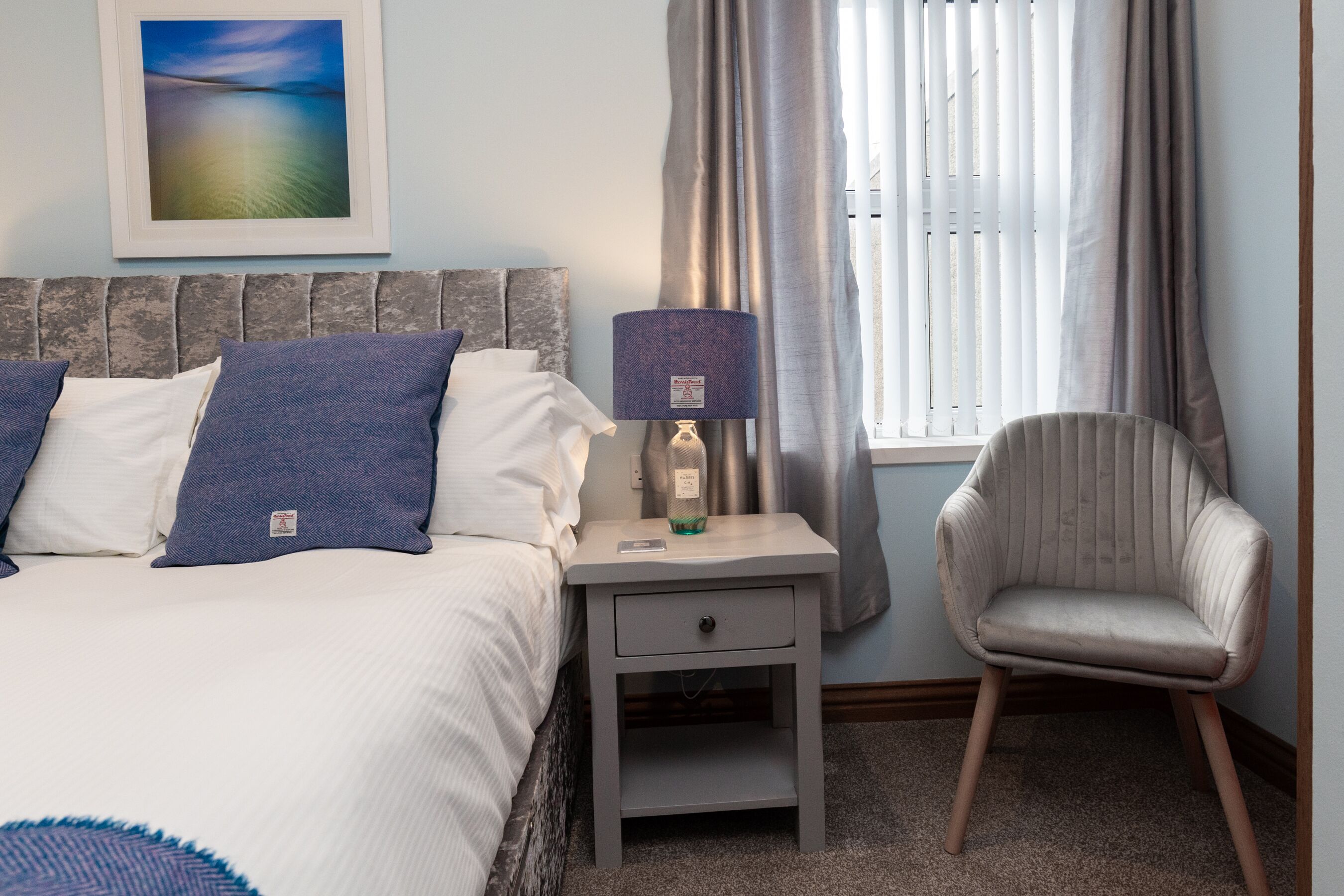 Een keurig opgemaakte slaapkamer met een bed, een nachtkastje met een lamp, een stoel en een raam met gordijnen.