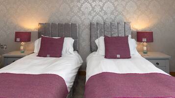 Een tweepersoonskamer met twee bedden, elk met hoofdbord, wit beddengoed en een paarse plaid. Er staan nachtkastjes met lampen en aan de muur een bloemenpatroon.