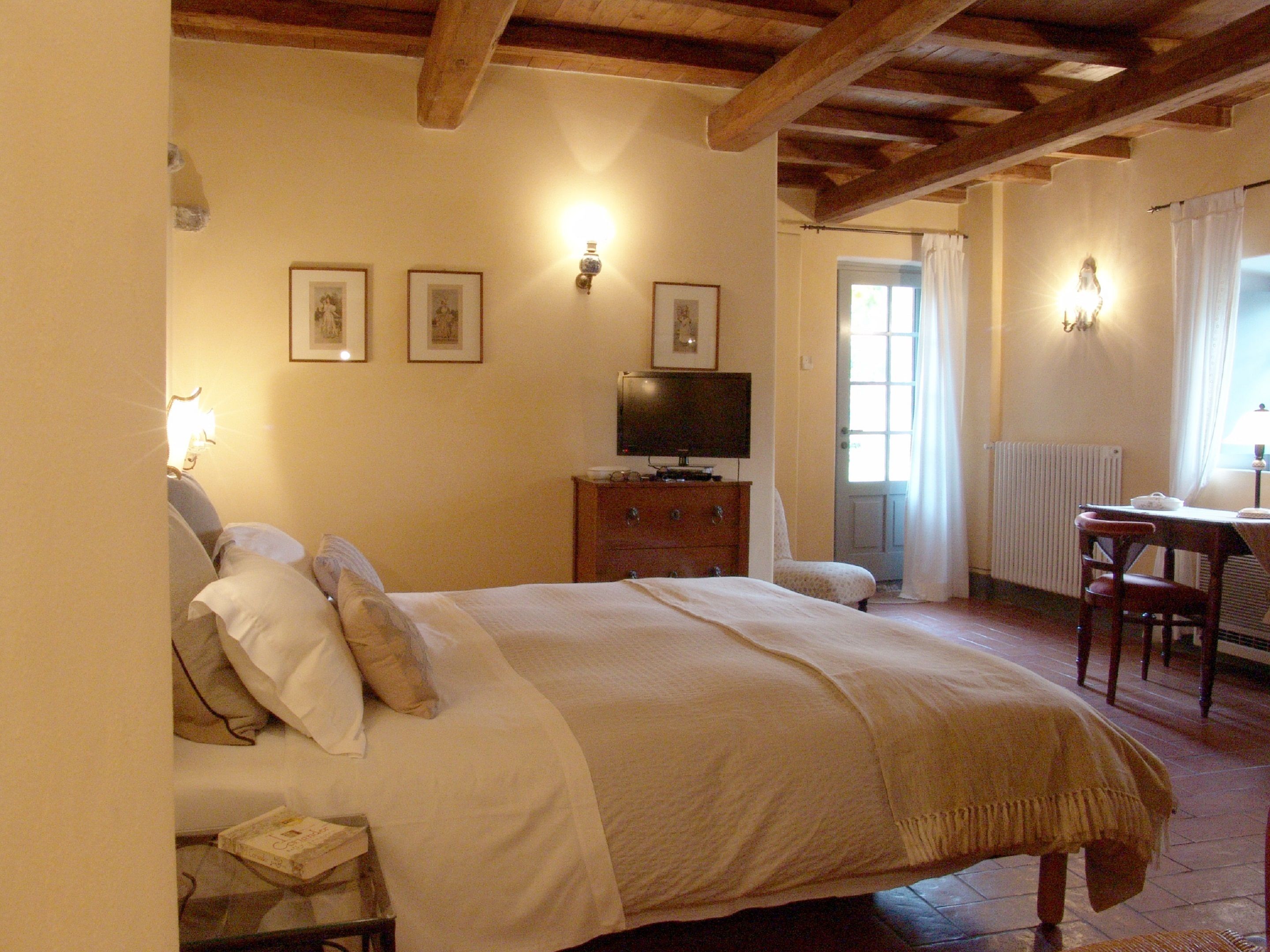 A bedroom with a bed, a television on a dresser, a desk with a chair, and a window with curtains.