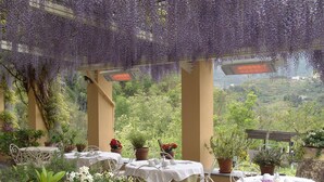 An outdoor dining area with tables set for two, surrounded by potted plants and a view of a hillside.