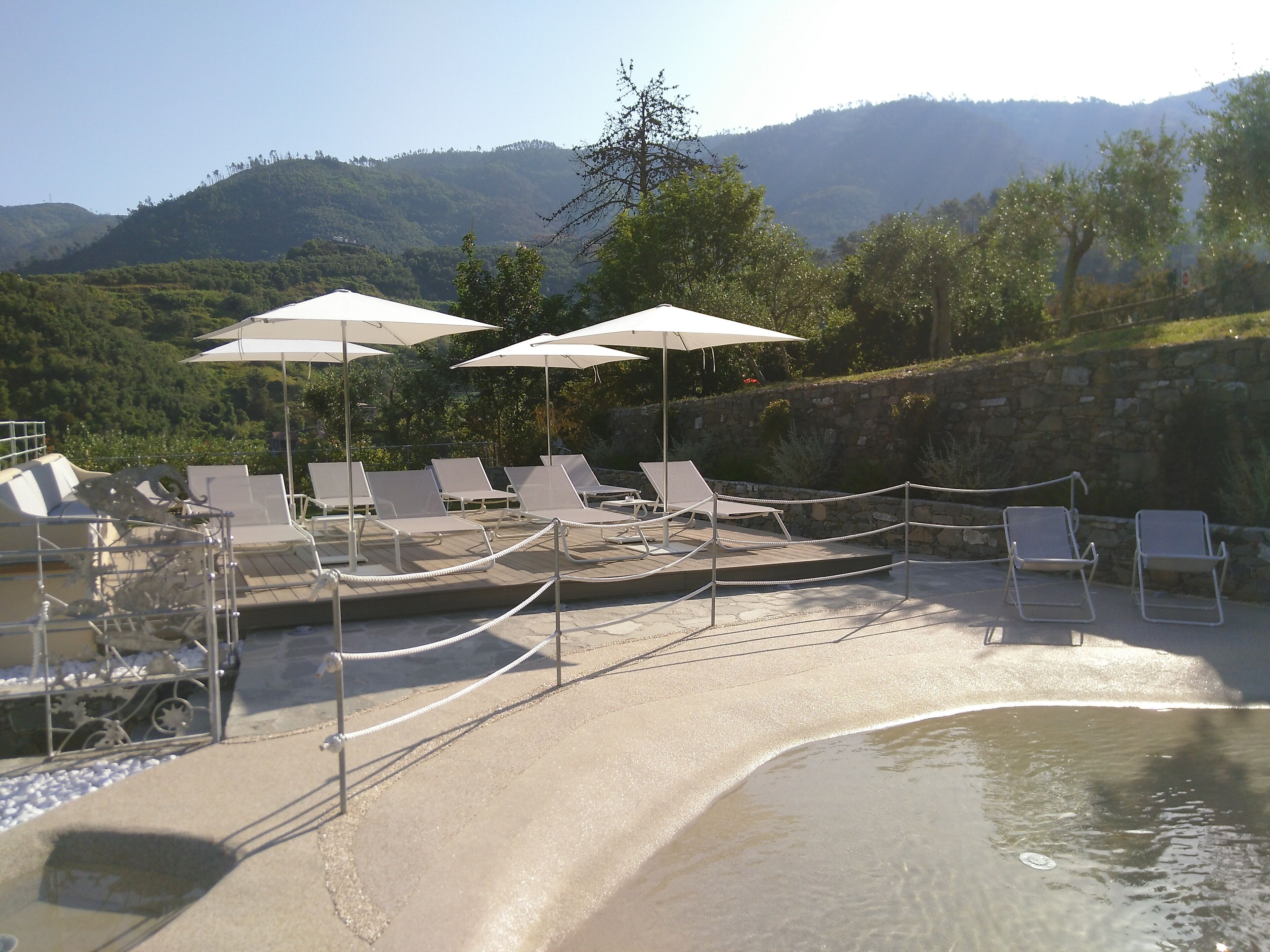 Un espace aménagé au bord de la piscine, avec des chaises longues et des parasols, entouré d’un mur de pierre et offrant une vue sur les montagnes.