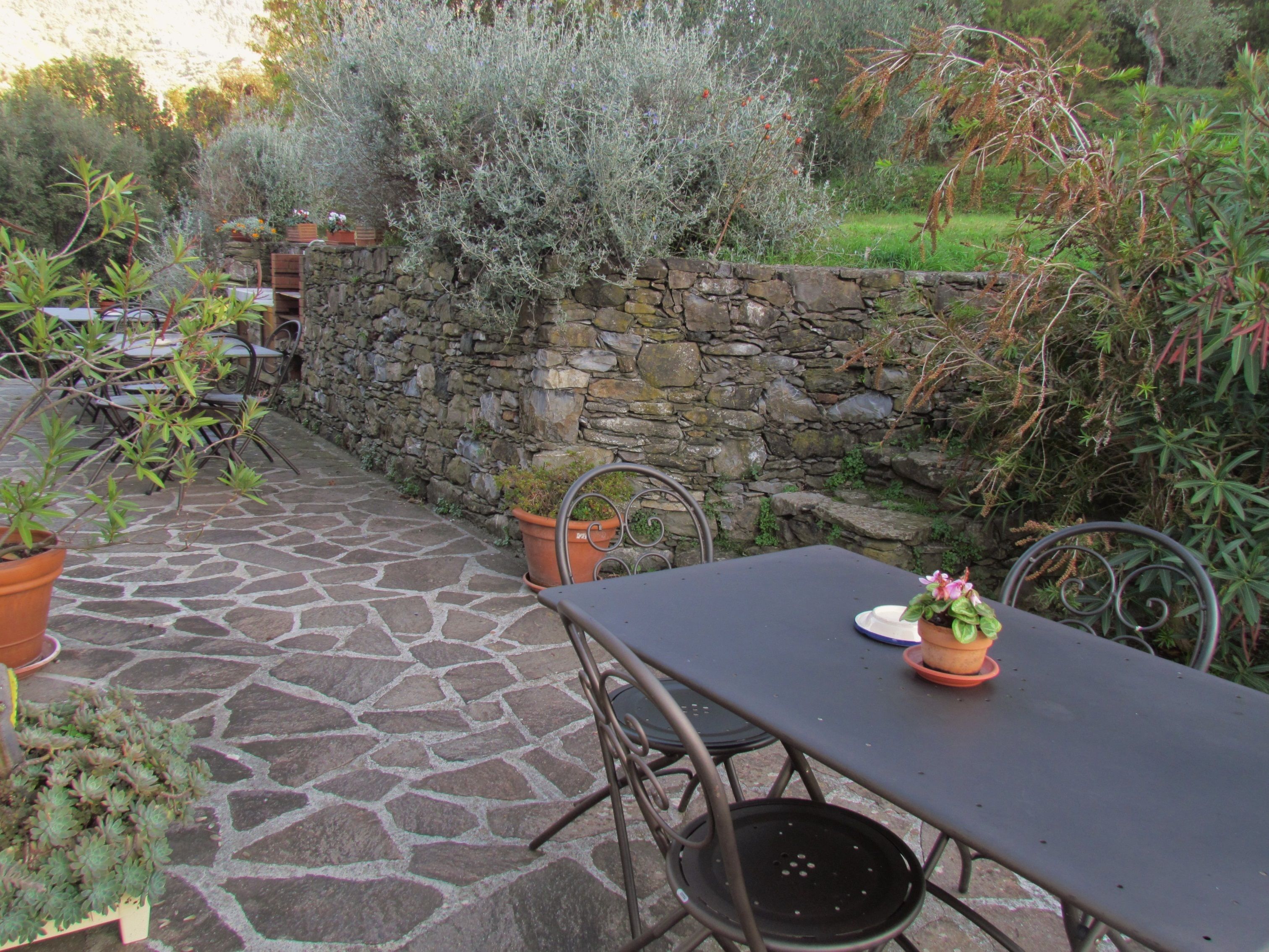 Un patio de piedra con una mesa y sillas, rodeado de macetas con plantas y un muro de piedra.