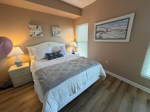 A bedroom with a bed, two bedside tables, a nightstand, a window with blinds, and a wall-mounted painting. - Enjoy Private Pools, Gulf Front Property and Spectacular Views at Crescent 312. (Destin)