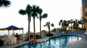 A swimming pool with palm trees, lounge chairs, and a beach view.