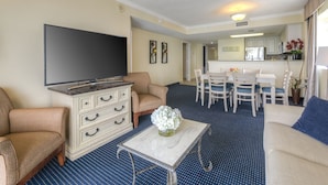 A living room with a flat-screen TV, a white coffee table with a vase of flowers, two beige sofas, and a dining area with a white table and chairs. - Gorgeous Remodeled 3BR, 2BA Camelot Oceanfront. In the heart of Myrtle Beach! (Myrtle Beach)