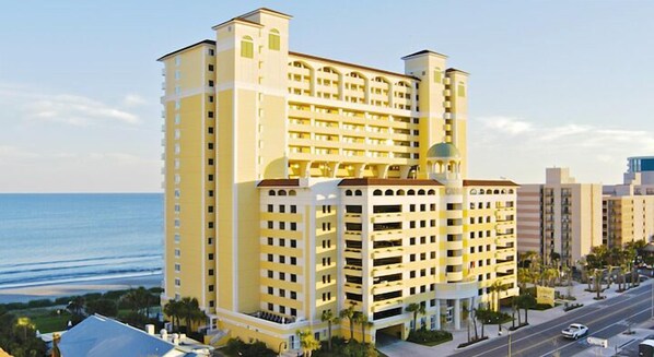 A multi-story hotel with a central courtyard, located near a beach with a clear view of the ocean.
