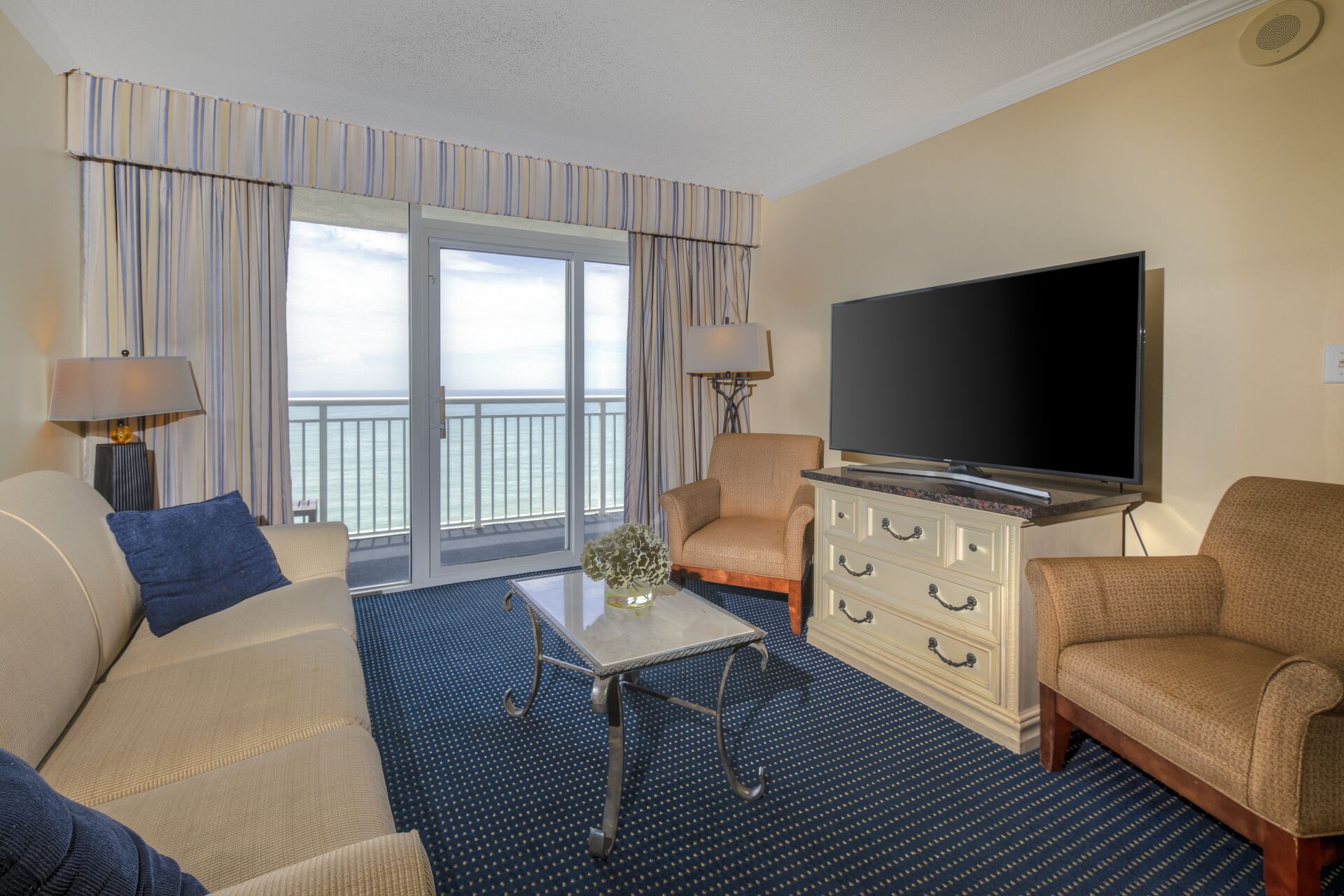 Una camera d'hotel con una grande TV, due poltrone, un divano, un tavolino da caffè e vista sull'oceano dalle finestre.