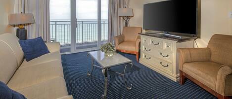 Una camera d'hotel con una grande TV, due poltrone, un divano, un tavolino da caffè e vista sull'oceano dalle finestre.