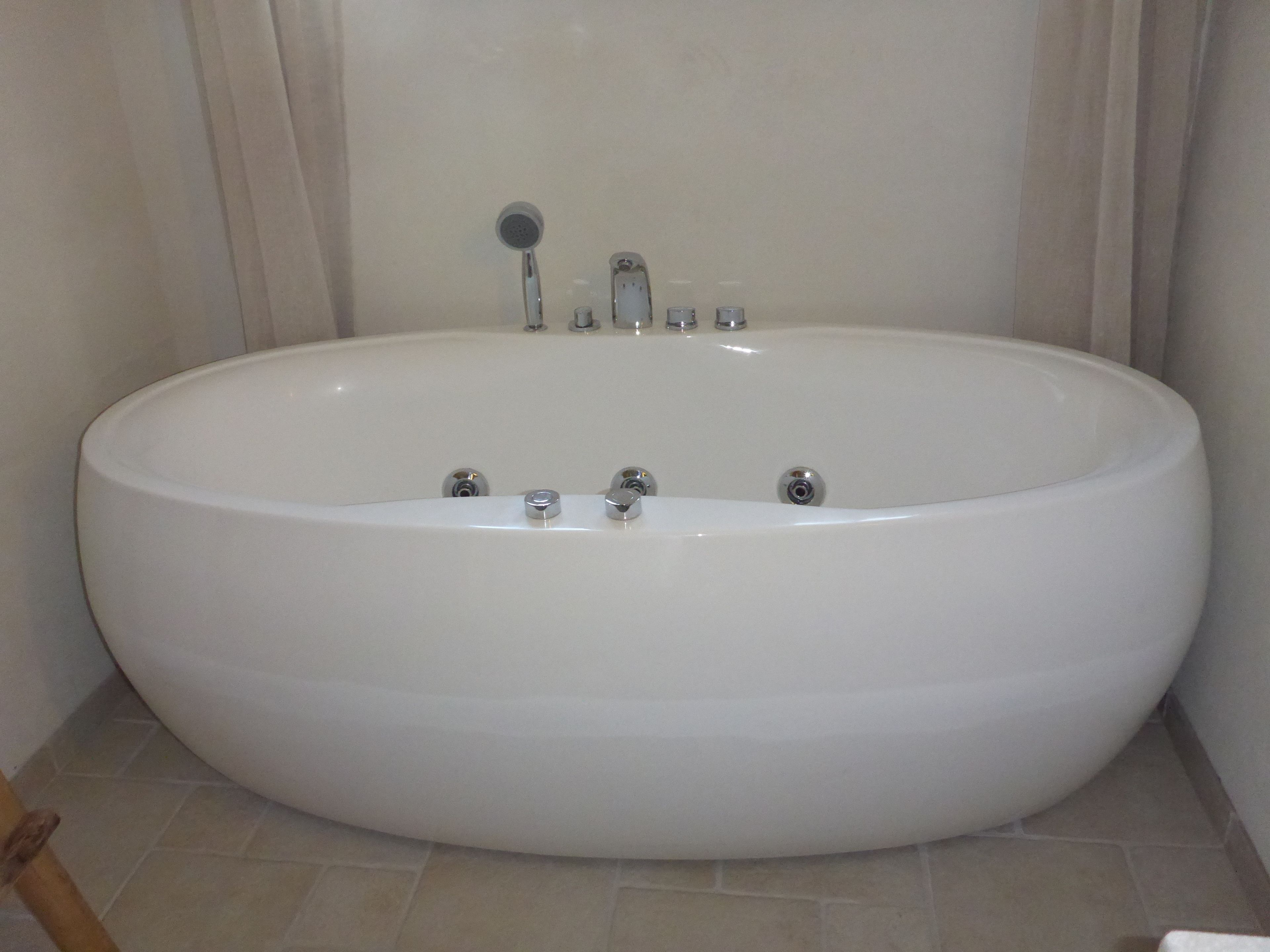 A large oval bathtub with multiple faucets and a handheld showerhead, set in a bathroom with tiled flooring.