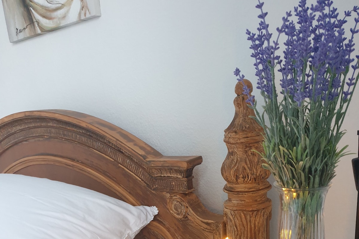 A bed with white linens, two lit candles on a wooden table, a vase with lavender, and a small statue.