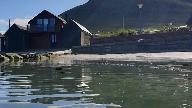 A coastal building with a wooden exterior and a balcony, situated near a body of water with a mountain in the background. - visitHOMES Faroe Islands (Leirvik)