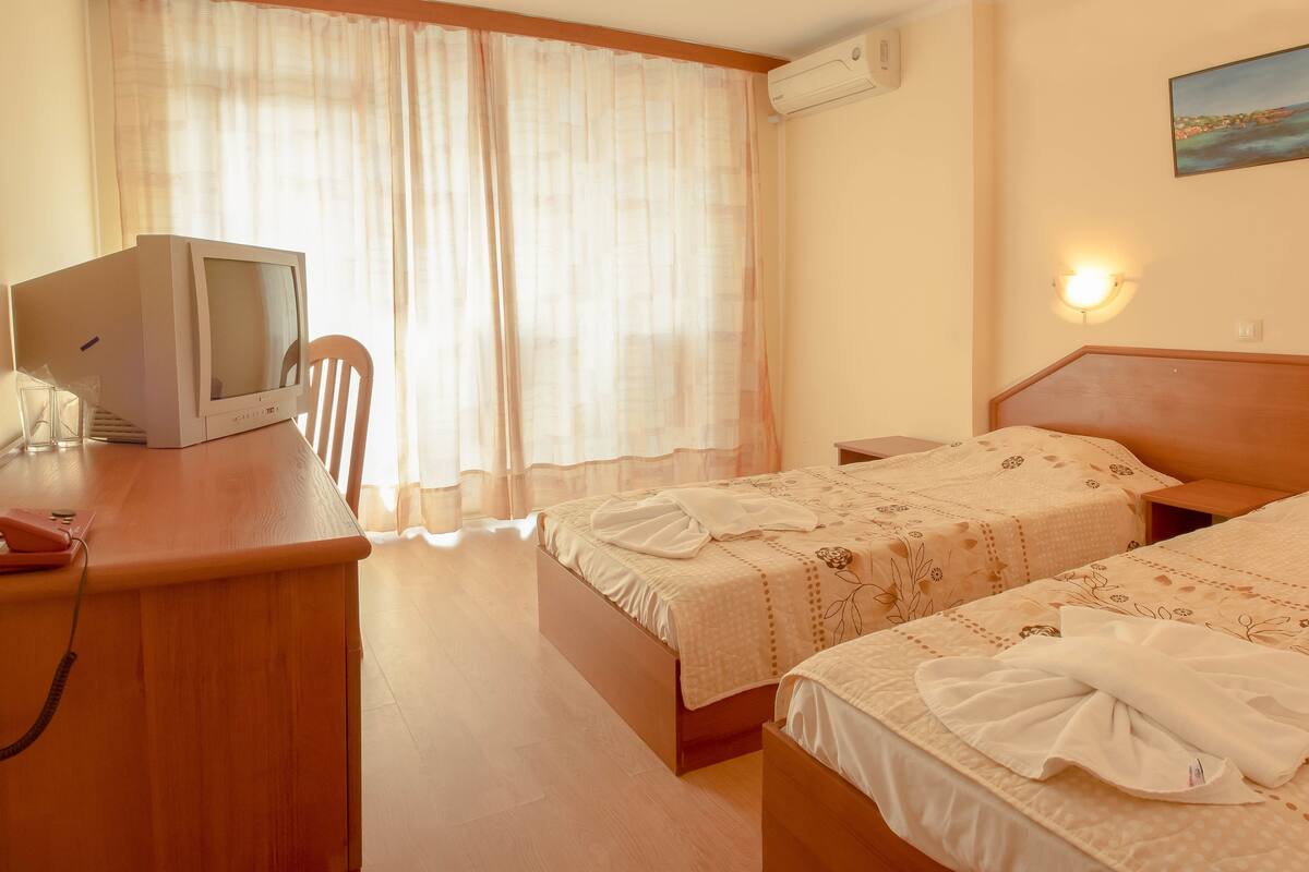Habitación de hotel con dos camas, un escritorio con teléfono, una silla, una ventana con cortinas translúcidas y un equipo de aire acondicionado.