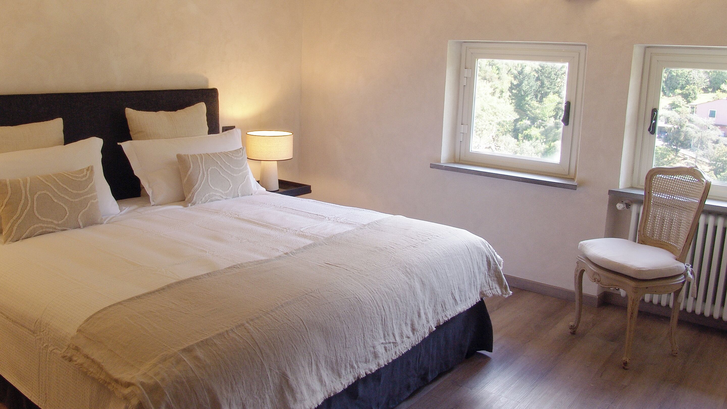 A bedroom with a large bed, a bedside table with a lamp, a chair, and a window with a view of trees.