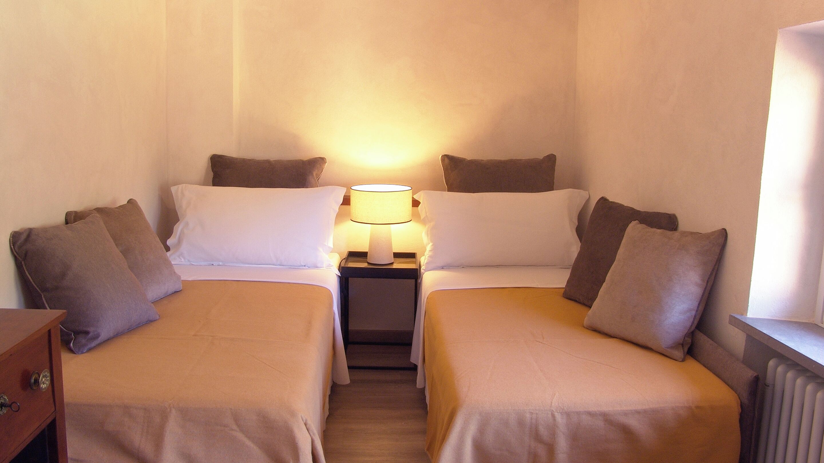 A small, cozy room with two single beds, each with a pillow and a bedside table with a lamp.