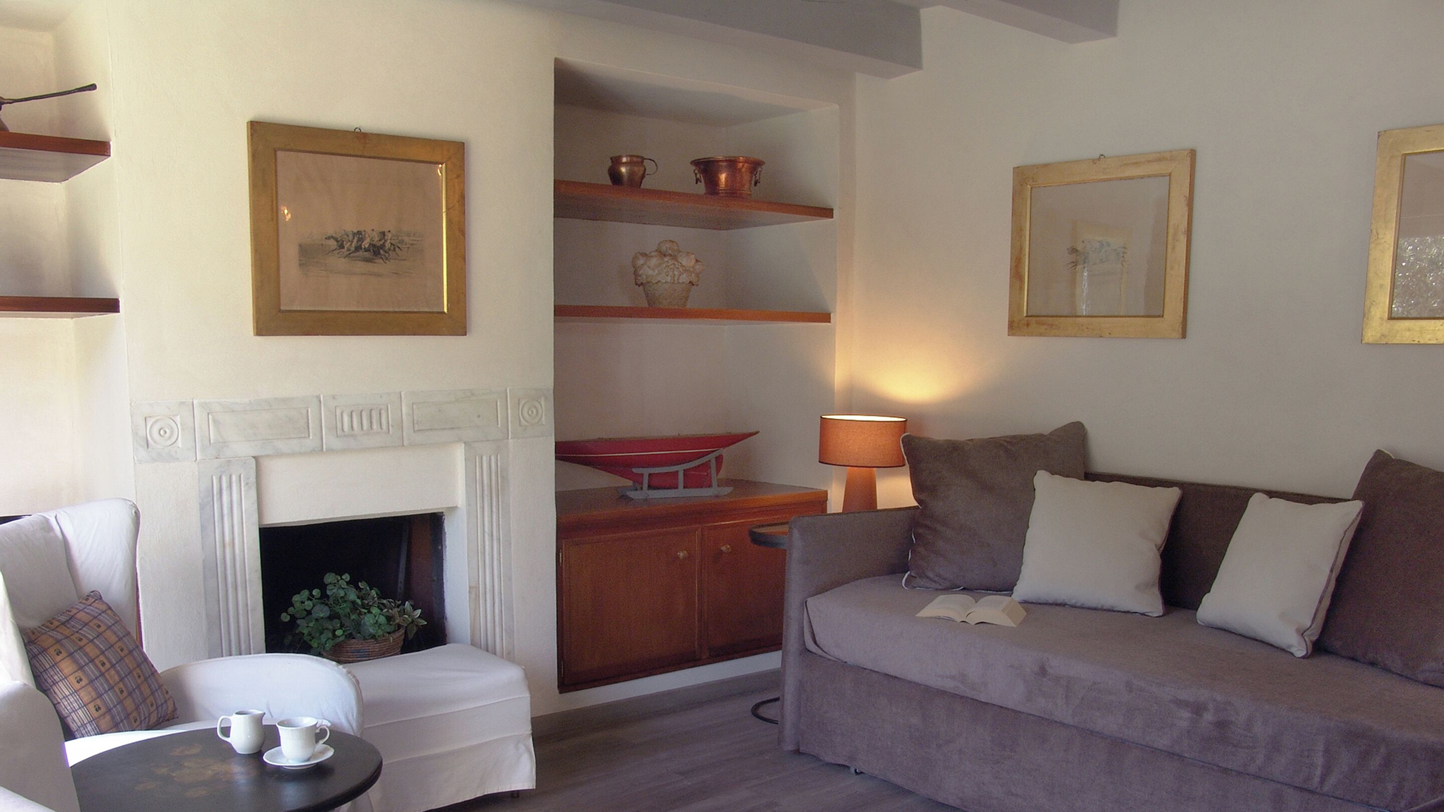 A living room with a fireplace, a sofa, a chair, a table with a cup, and framed pictures on the wall.