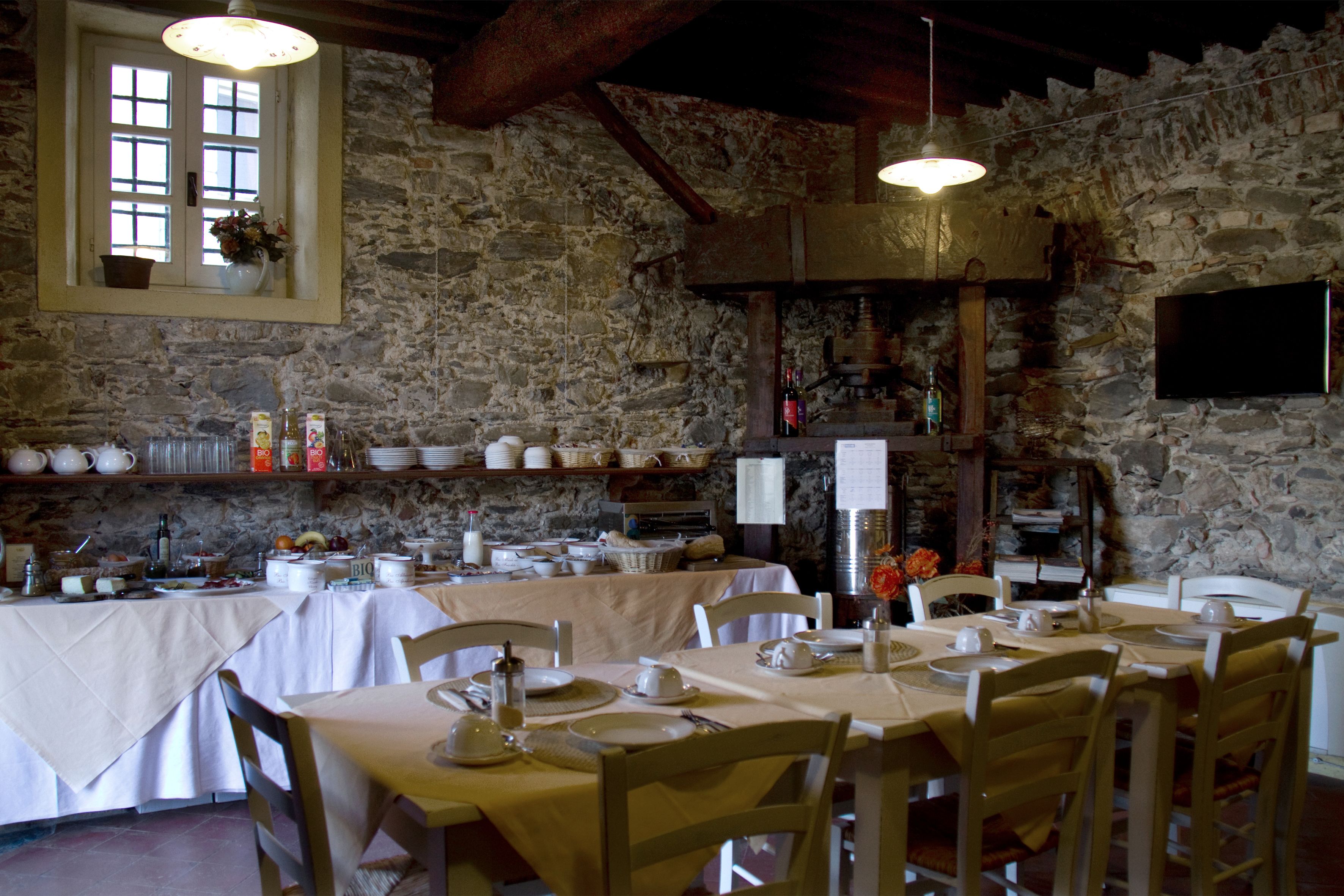 A rustic dining area with stone walls, wooden beams, and a large window. Tables are set with white tablecloths and chairs.
