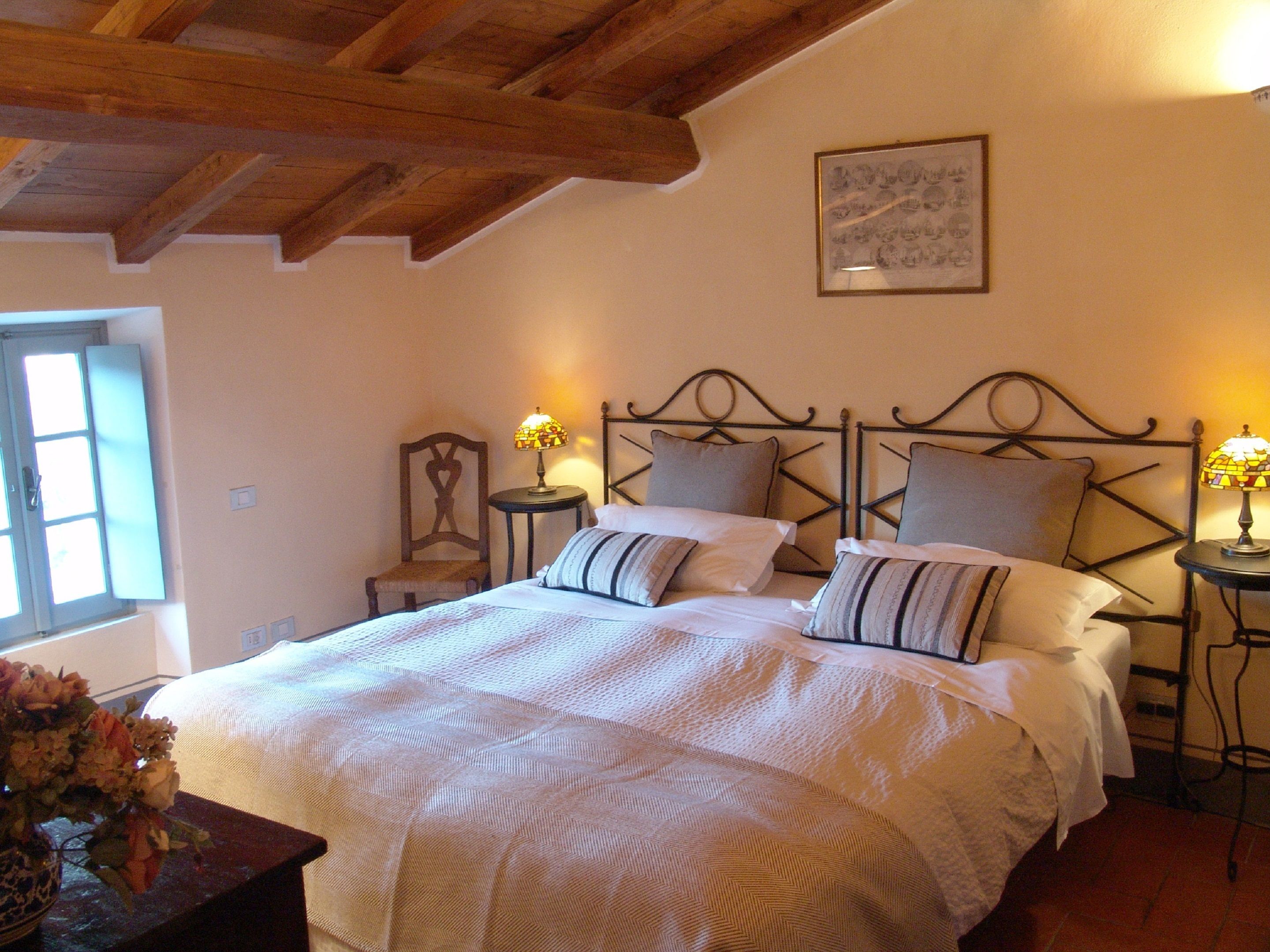 A bedroom with a wooden ceiling, a bed with white bedding, two bedside tables with lamps, and a window with a view of the outside.