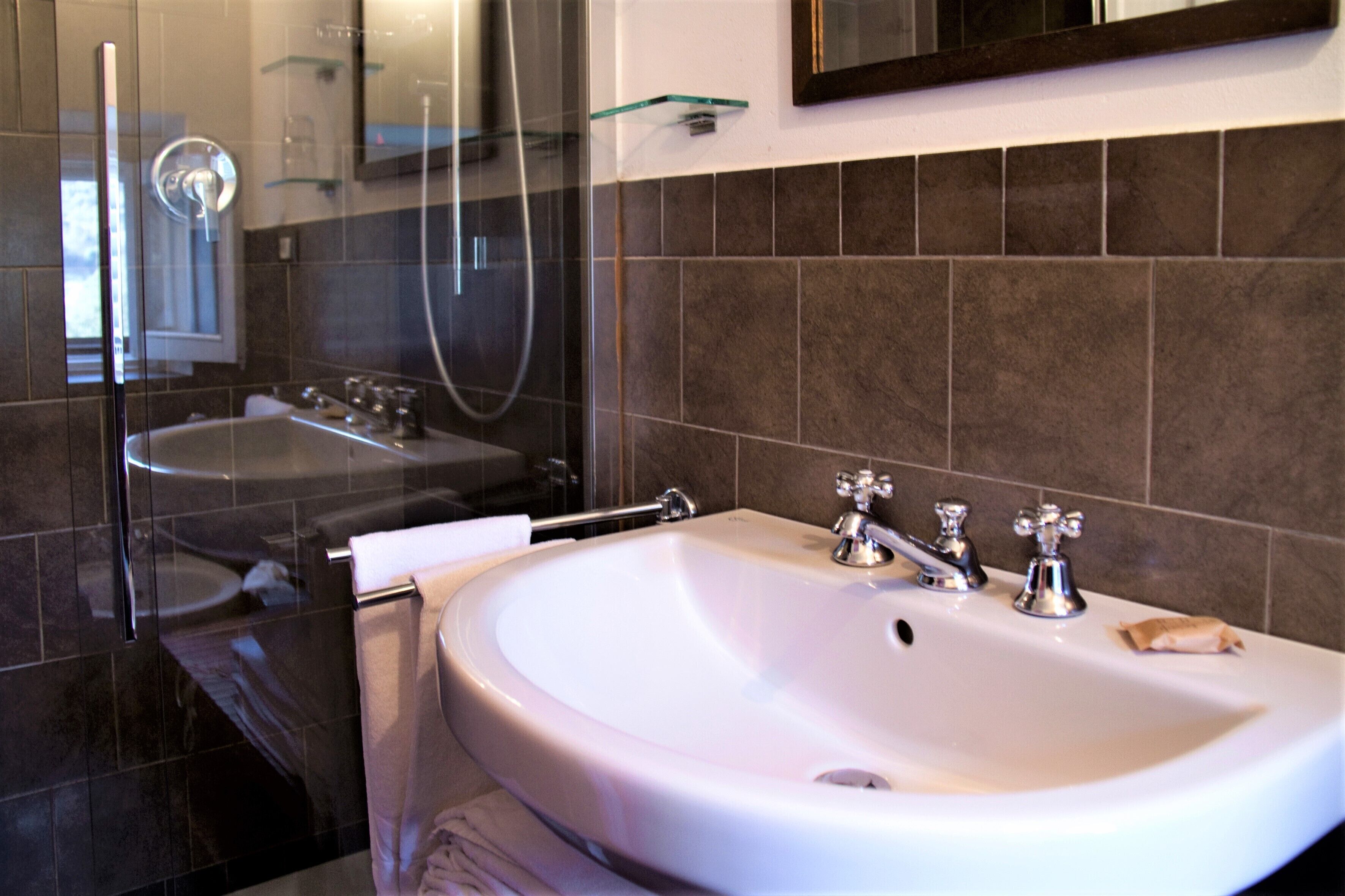 Ein modernes Badezimmer mit einem weißen Waschbecken, zwei Wasserhähnen und einer Duschkabine aus Glas.