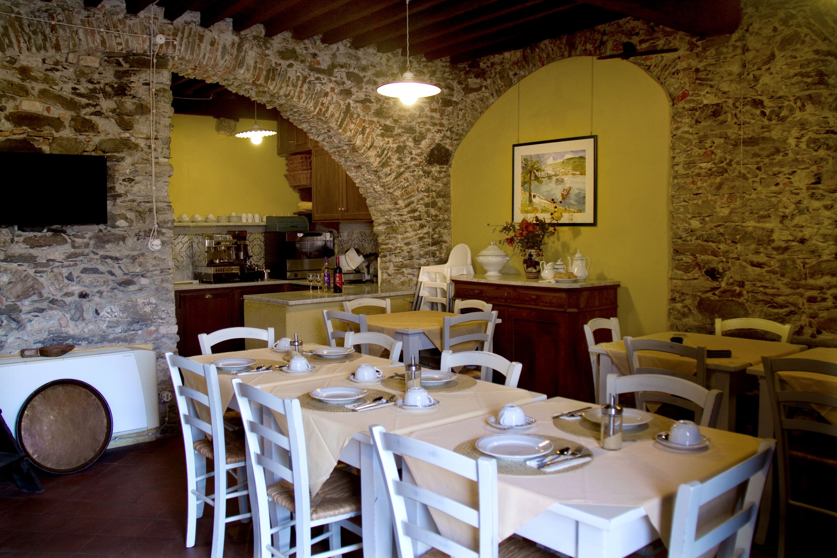 Un comedor con paredes de piedra, mobiliario blanco y un arco de piedra. Las mesas están preparadas con platos, tazas y cubiertos.