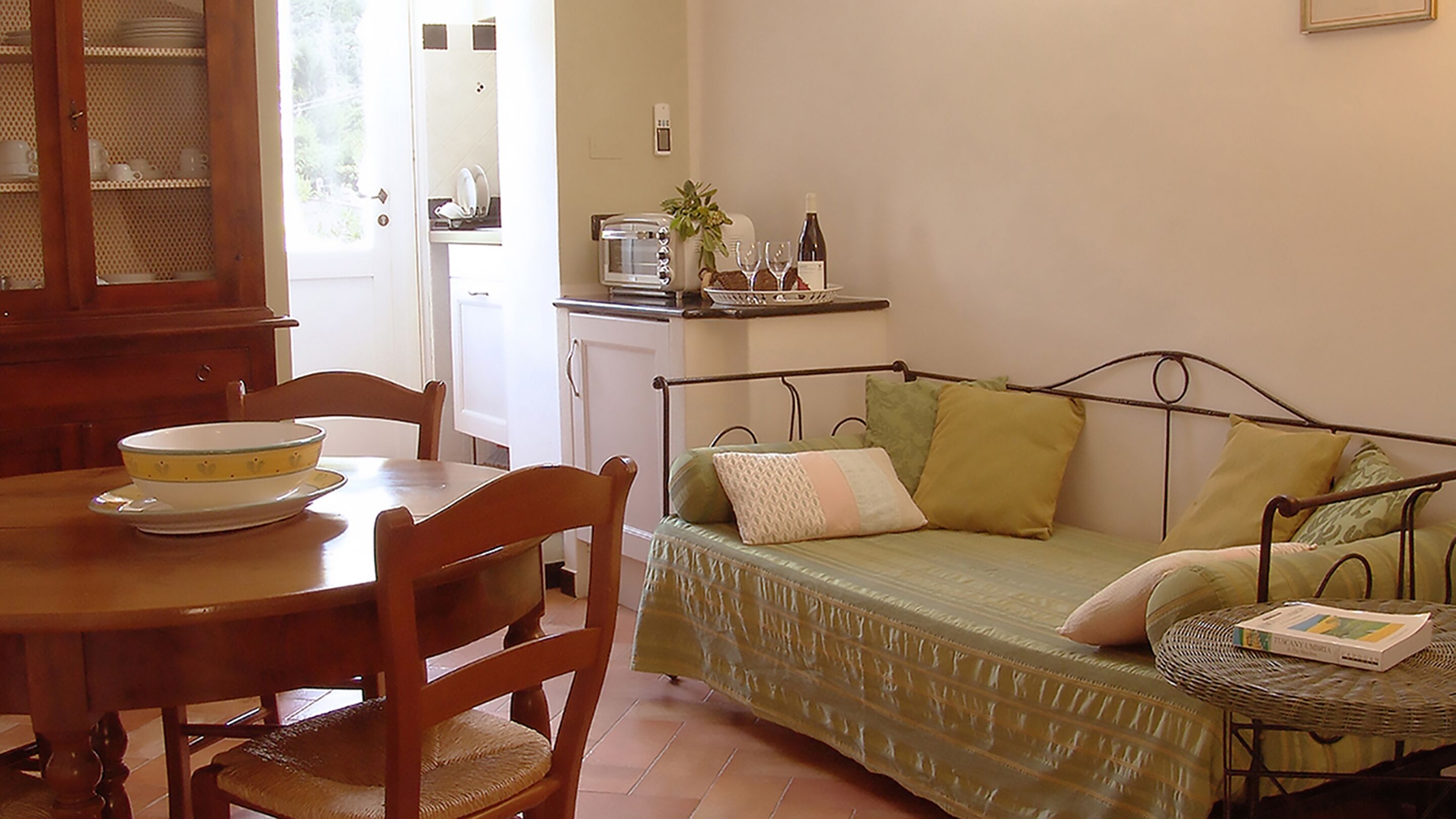 Una habitación acogedora con una mesa de comedor, sillas, una cama con almohadas, una mesita con un libro y una zona de cocina con un tostador y una planta.