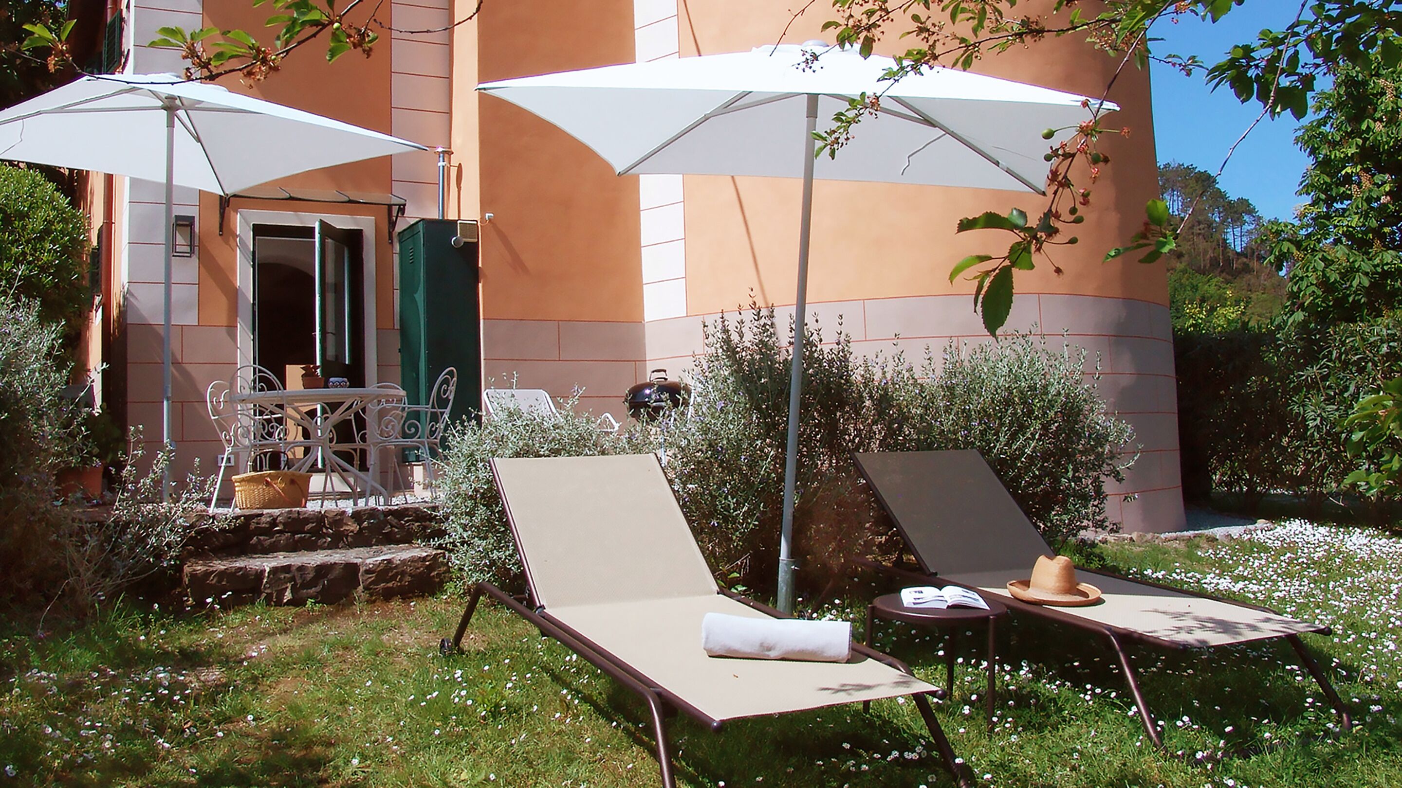 Un espace extérieur aménagé avec deux fauteuils et des parasols, une petite table sur laquelle se trouvent un chapeau et un livre, et un bâtiment avec une porte verte et des fenêtres en arrière-plan.
