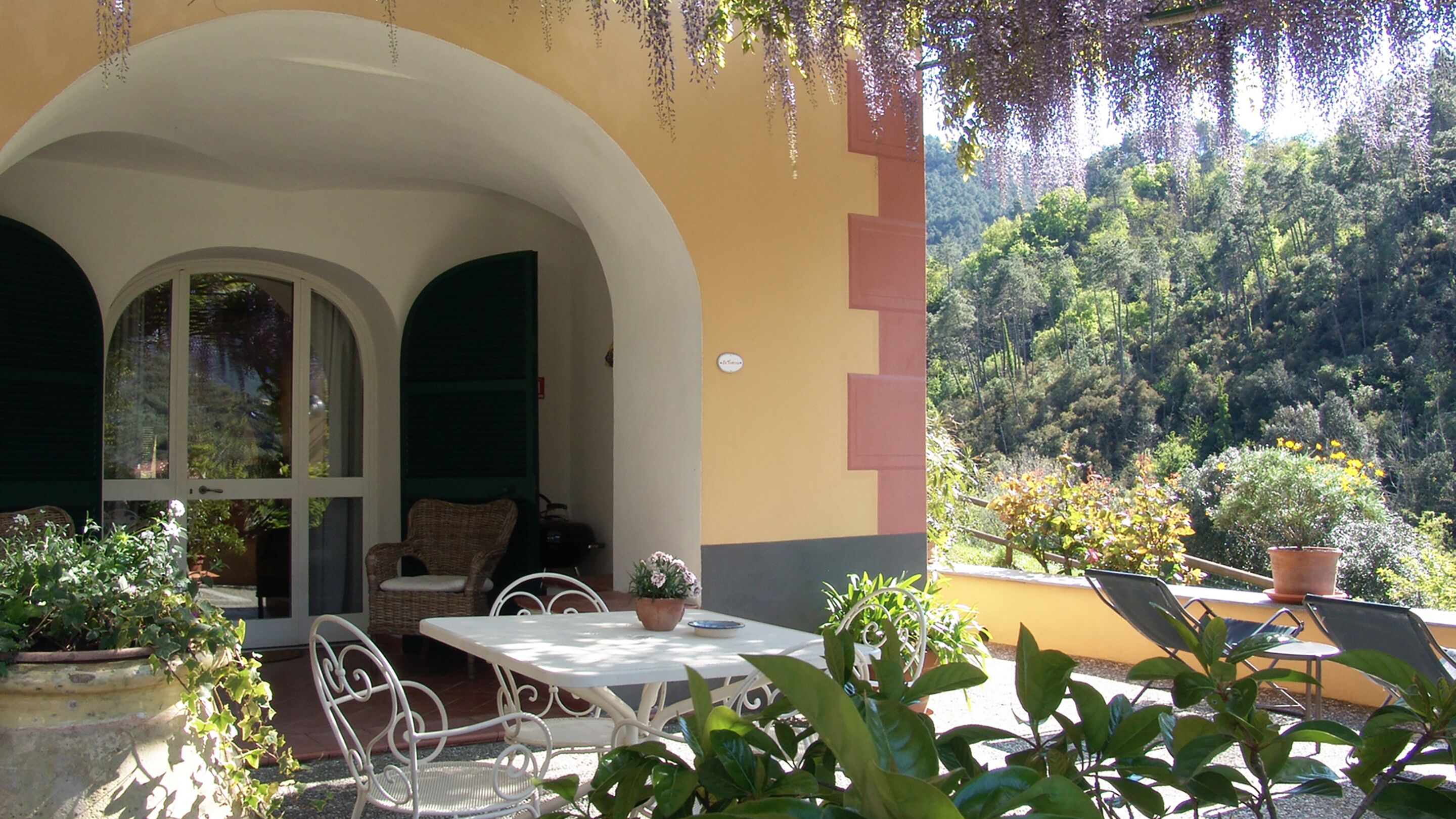 A patio with a table and chairs, surrounded by potted plants and a view of a forested hillside.