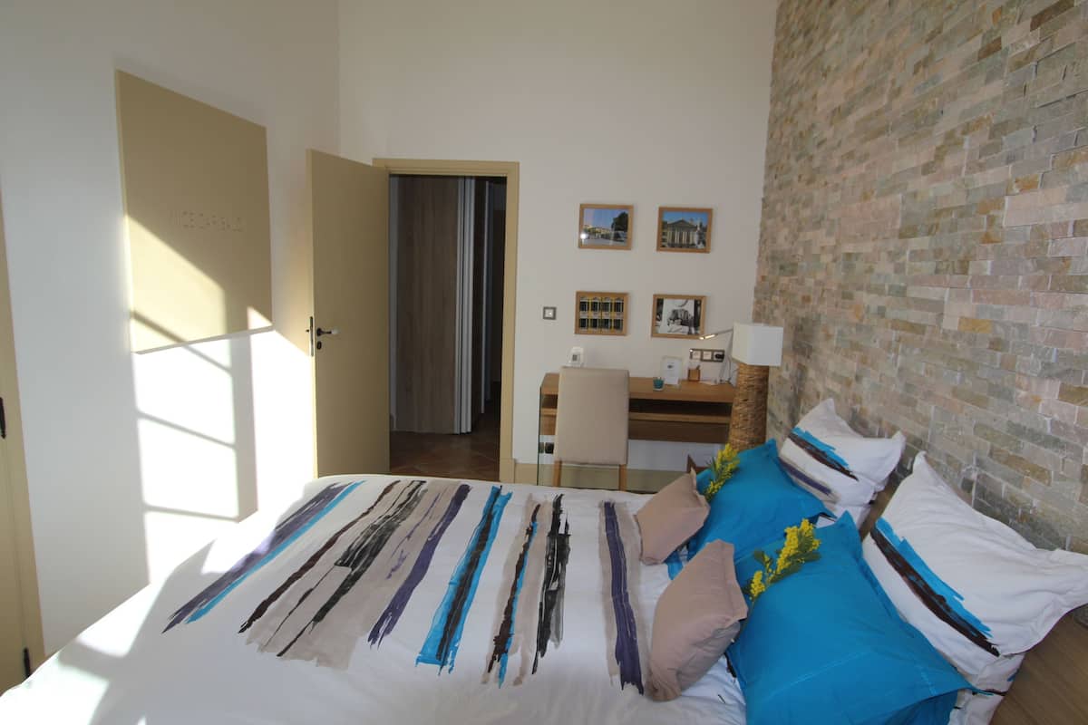 A bedroom with a bed, a desk, and a chair. There are pictures on the wall and a brick wall on one side.