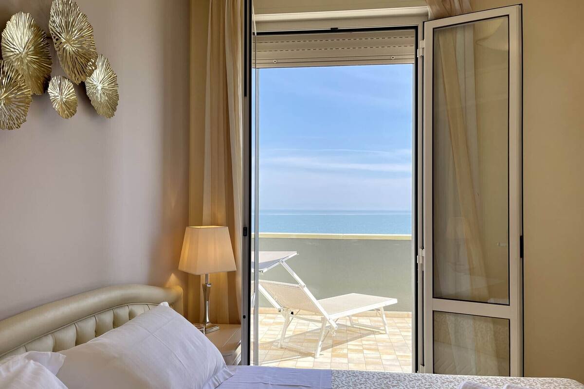 Une chambre d’hôtel avec un lit, une lampe, une chaise et une vue sur la mer, visible par une porte ouverte.