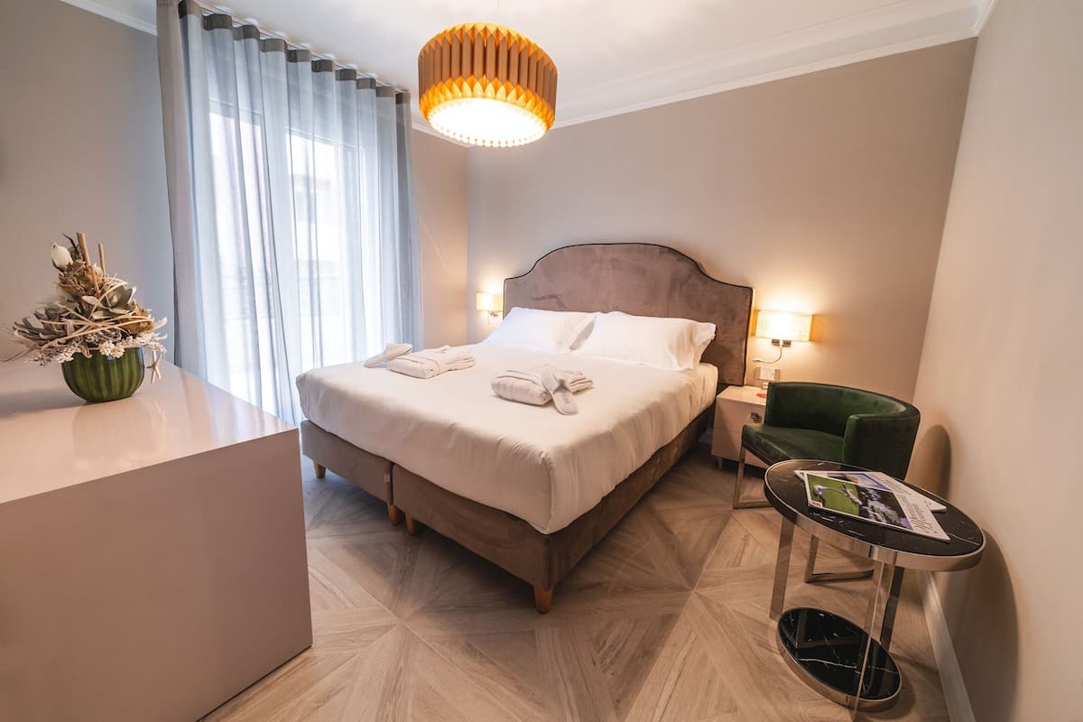 Une chambre d’hôtel avec un lit, une table de chevet, un fauteuil vert, une petite table avec un magazine et une plante décorative.