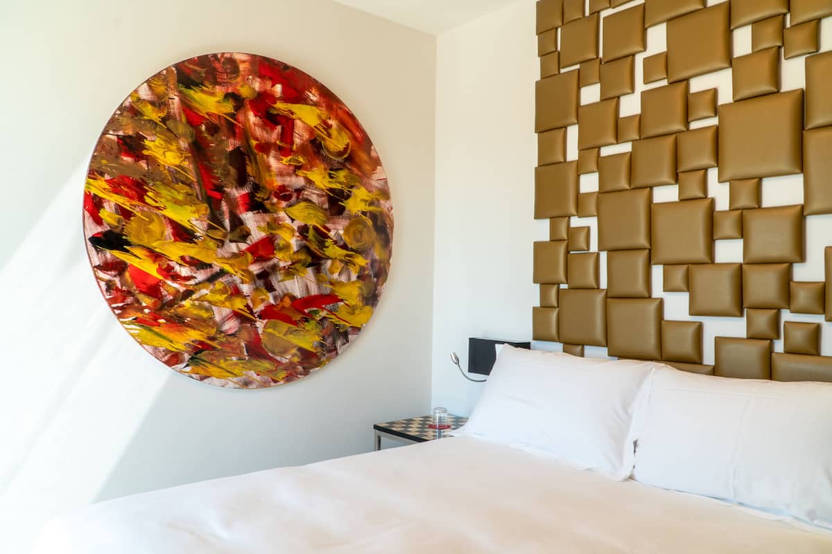 Une chambre d’hôtel moderne avec un grand tableau abstrait au mur, une tête de lit aux motifs géométriques et un lit parfaitement fait.