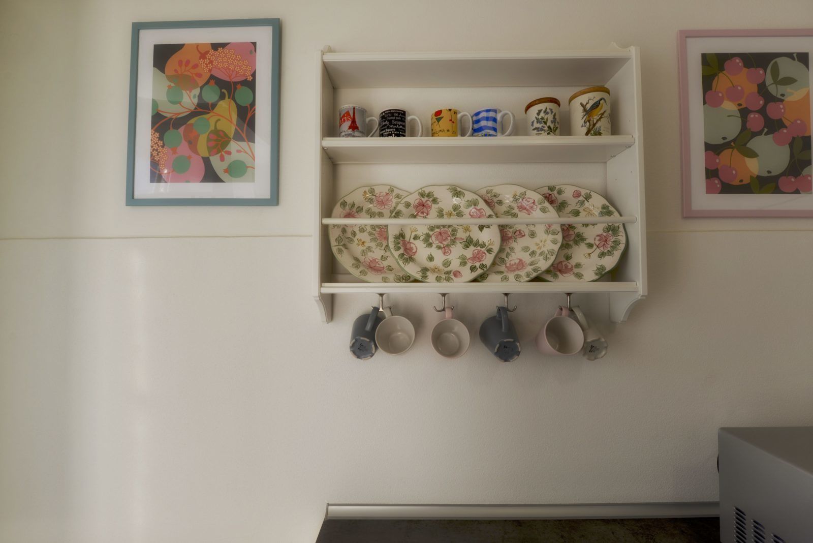 A kitchen wall with a white shelf holding plates, mugs, and a framed floral painting.