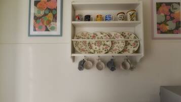 Un mur de cuisine avec une étagère blanche où sont posées des assiettes, des tasses et un tableau floral encadré.