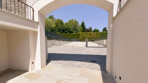 Ein bogenförmiger Eingang führt zu einem gepflasterten Innenhof mit einem Metallgitter und einer Hecke im Hintergrund.