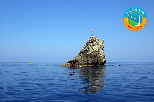A rocky island with a small hut and a flag, surrounded by calm blue waters. - EcoBelmonte Albergo Diffuso (Belmonte Calabro)