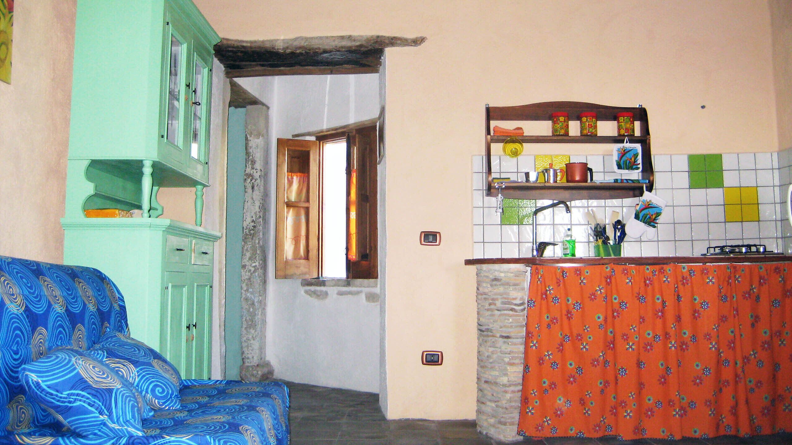 Una habitación con sofá verde, zona de cocina con salpicadero de azulejos y ventana con cortinas.