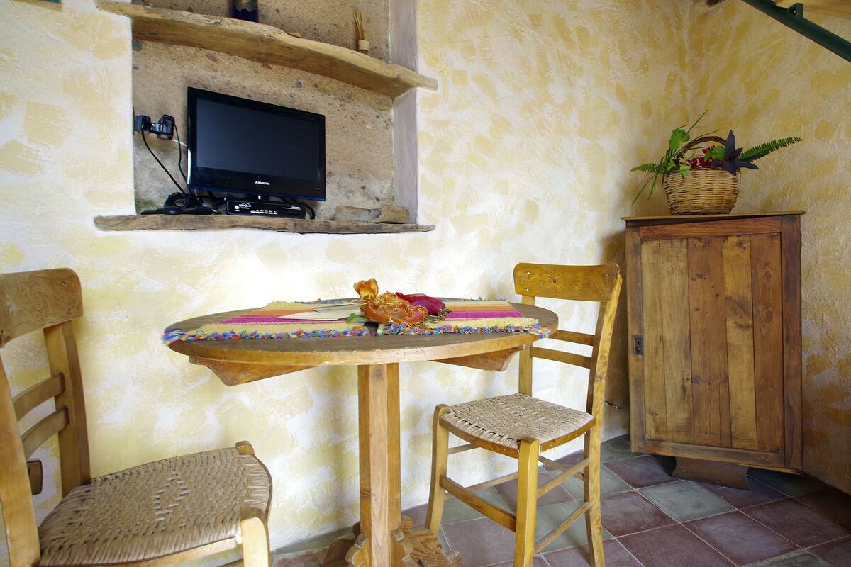 A rustic room with a round wooden table, two chairs, a television mounted on a wall, a wooden cabinet, and a wicker basket with plants.